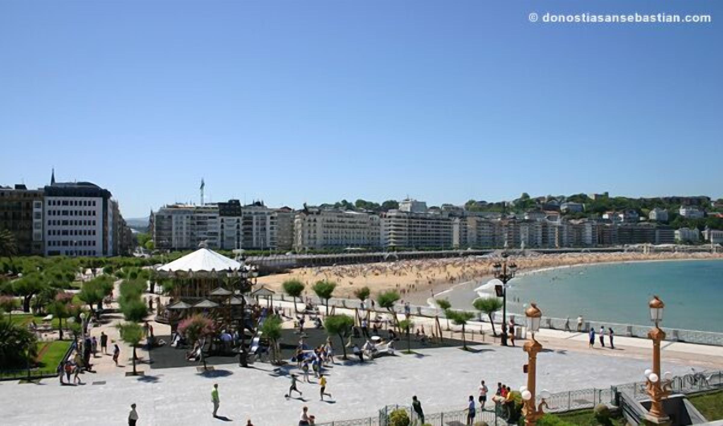Un paseo marítimo con playa, un quiosco y una hilera de edificios al fondo.