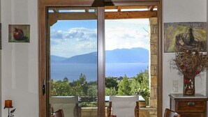 Un coin repas avec une table dressée pour deux personnes, une vue sur la mer et les montagnes, et un vase rempli de fleurs séchées posé sur une commode.