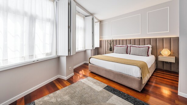 A bedroom with a bed, a nightstand, a lamp, a window with curtains, and a wooden floor. - Victoria Project - Apartment I - Private Parking (Porto)