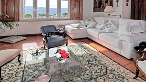 Un salon avec un canapé blanc, un fauteuil bleu, une table basse en verre ornée d’un vase de fleurs, un tapis à motifs et une vue sur les montagnes, visible à travers de grandes fenêtres.
