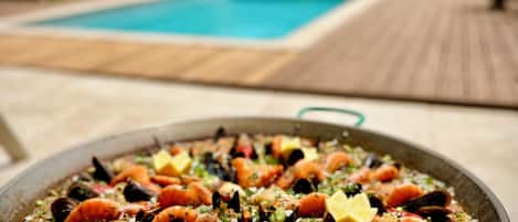 A seafood paella on a table with a pool and greenery in the background.