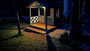 Ein kleines Holzhaus mit einer Veranda und einem eingezäunten Eingang, umgeben von Bäumen und einem Kiesweg.