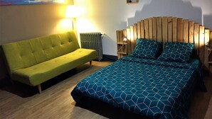Ein Zimmer mit einem grünen Sofa, einem Bett mit einem geometrisch gemusterten Überwurf, einem hölzernen Kopfteil, einer Wandlampe und einem kleinen Bilderrahmen.