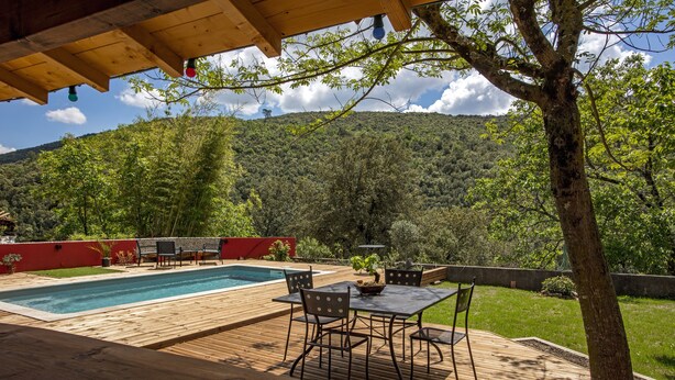 Eine Holzterrasse mit Pool, Gartenmöbeln und Blick auf einen bewaldeten Hügel.