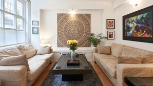 Una sala de estar moderna con dos sofás color beige, una mesa de centro de vidrio y un tapiz grande en la pared.