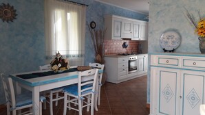A kitchen with white cabinets, a dining table with chairs, and a window with curtains. - The Lapwings (Calasetta)