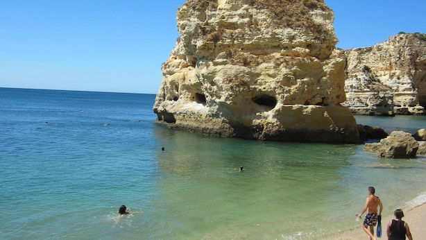 A beach with clear turquoise water, a large rock formation, and people walking along the shore.