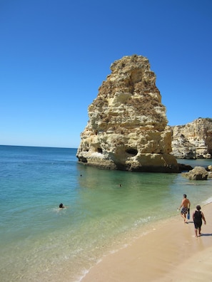 Uma praia com águas turquesa cristalinas, uma grande formação rochosa e pessoas caminhando pela orla.