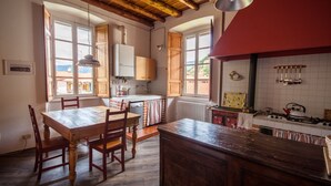 A rustic kitchen with wooden beams, a dining table with chairs, a red ceiling, and a view of the outside through windows.