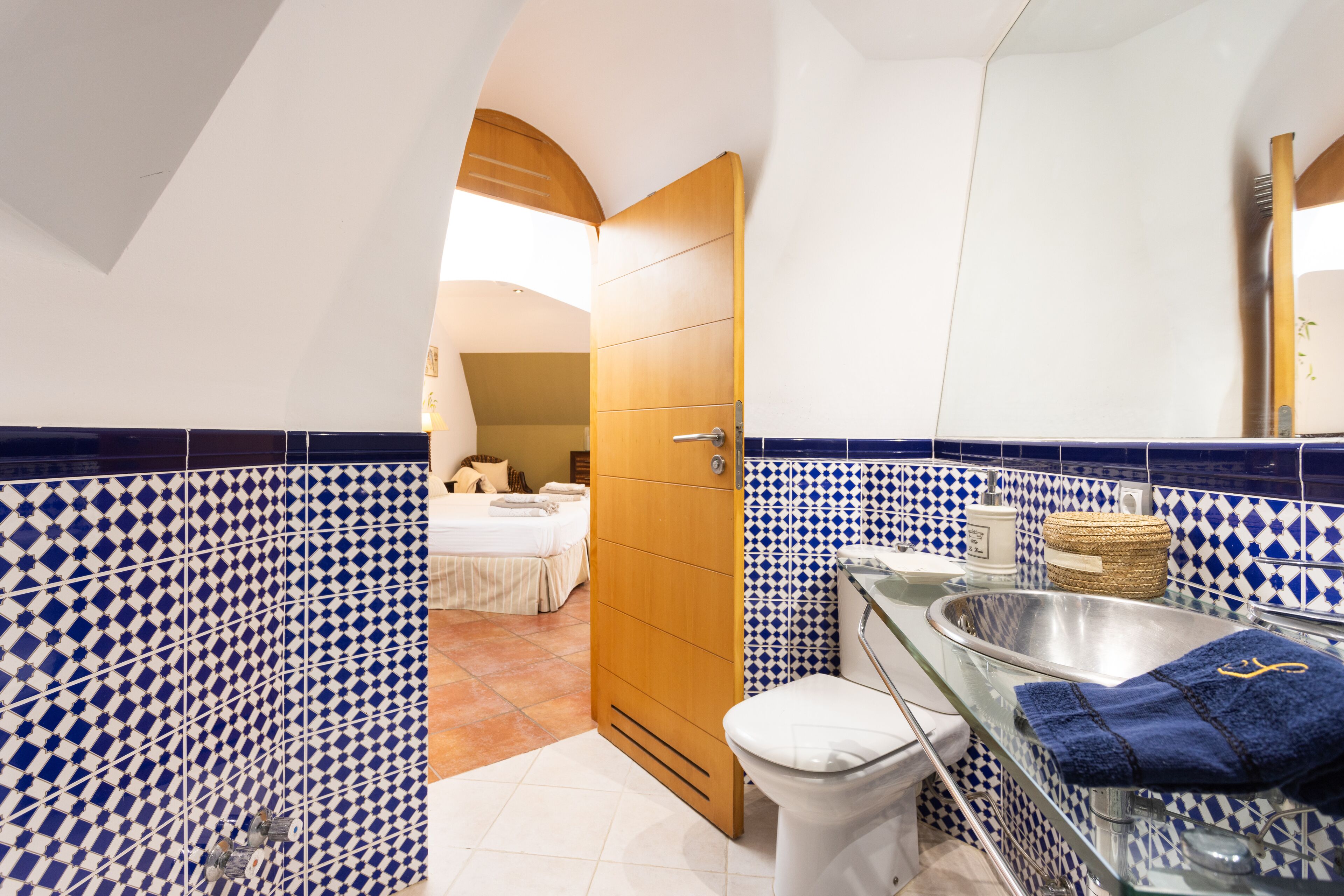 Une salle de bain avec un mur recouvert de carreaux bleus et blancs, une douche en verre, des toilettes blanches et une porte en bois donnant sur une chambre.
