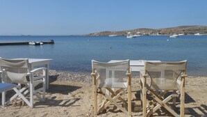 A beach with two white chairs and a table, a calm sea, boats, and a distant hill.