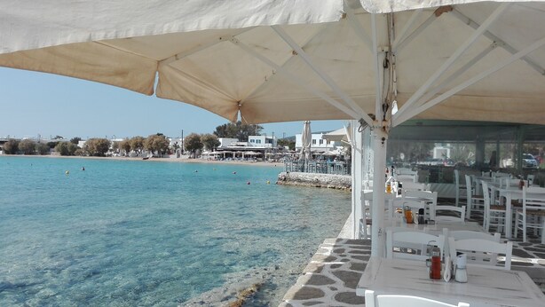 A seaside restaurant with outdoor seating, clear blue waters, and a view of the beach and buildings in the background.