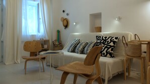 A living room with a white sofa, wooden chairs, a small table, and a window with white curtains.