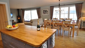 A spacious living area with a wooden dining table, chairs, and a large window. A kitchen area with a wooden countertop and a bowl of fruit is visible. - Exceptional House In Superb Location in NTS Conservation Area, Stunning Sea View (Kyle of Lochalsh)