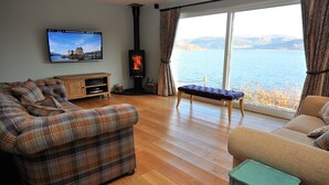 A living room with a plaid sofa, a bench, a TV, and a wood-burning stove with a view of a lake. - Exceptional House In Superb Location in NTS Conservation Area, Stunning Sea View (Kyle of Lochalsh)