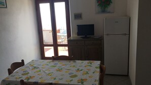 Un comedor con una mesa cubierta con un mantel estampado de frutas, sillas de madera, un refrigerador, un televisor sobre un mueble de madera y una puerta que da al exterior.