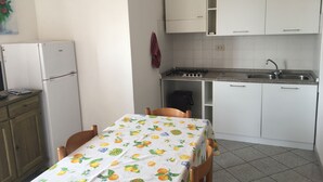 Una cocina compacta con armarios blancos, una estufa a gas y un lavamanos. Hay una mesa con mantel estampado de frutas y dos sillas.