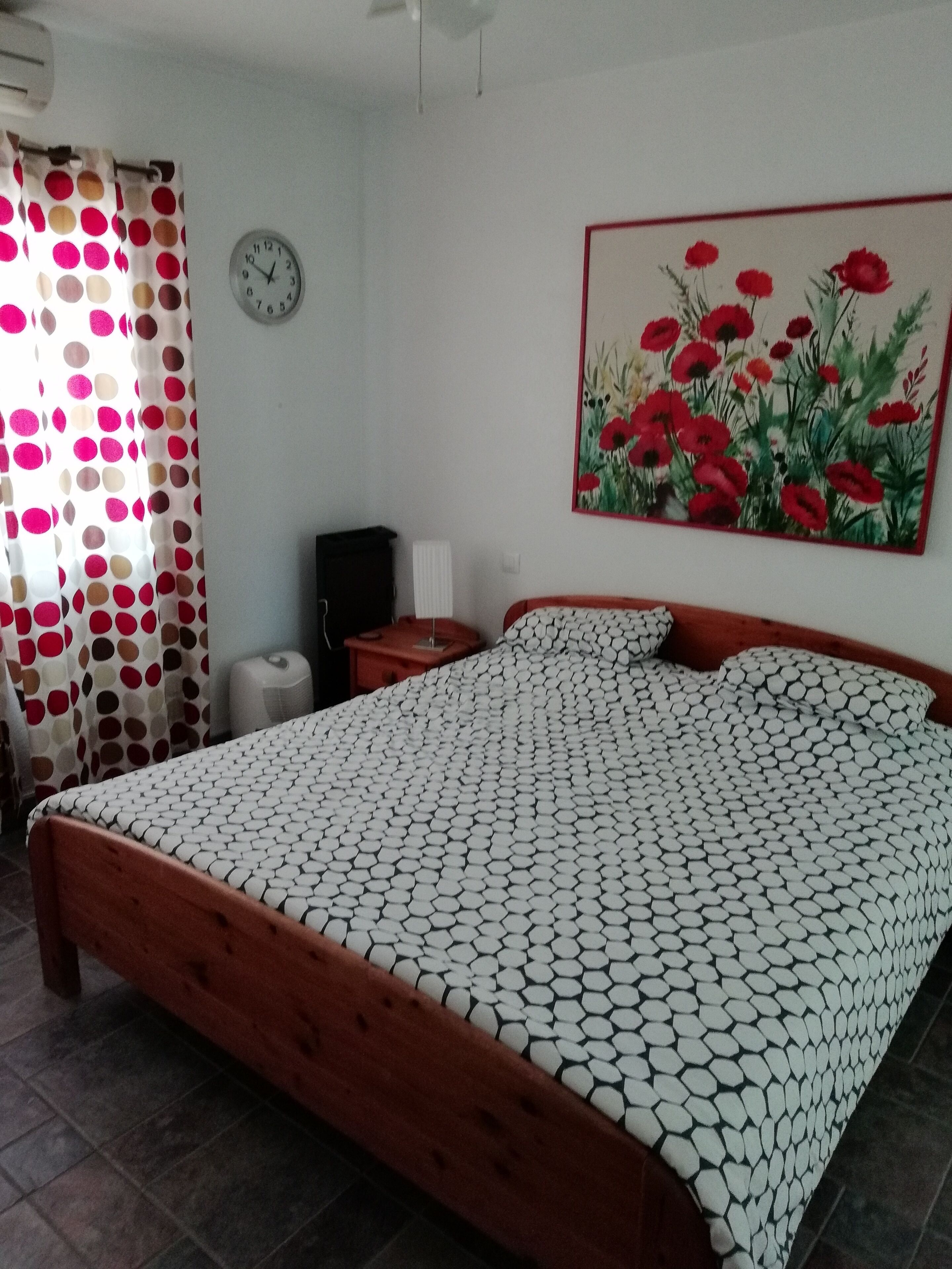 A bedroom with a bed, a clock, a radiator, and a painting of flowers.