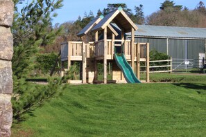 Children can play on the multi-level play structure with a green slide in a natural setting surrounded by trees and shrubs.
