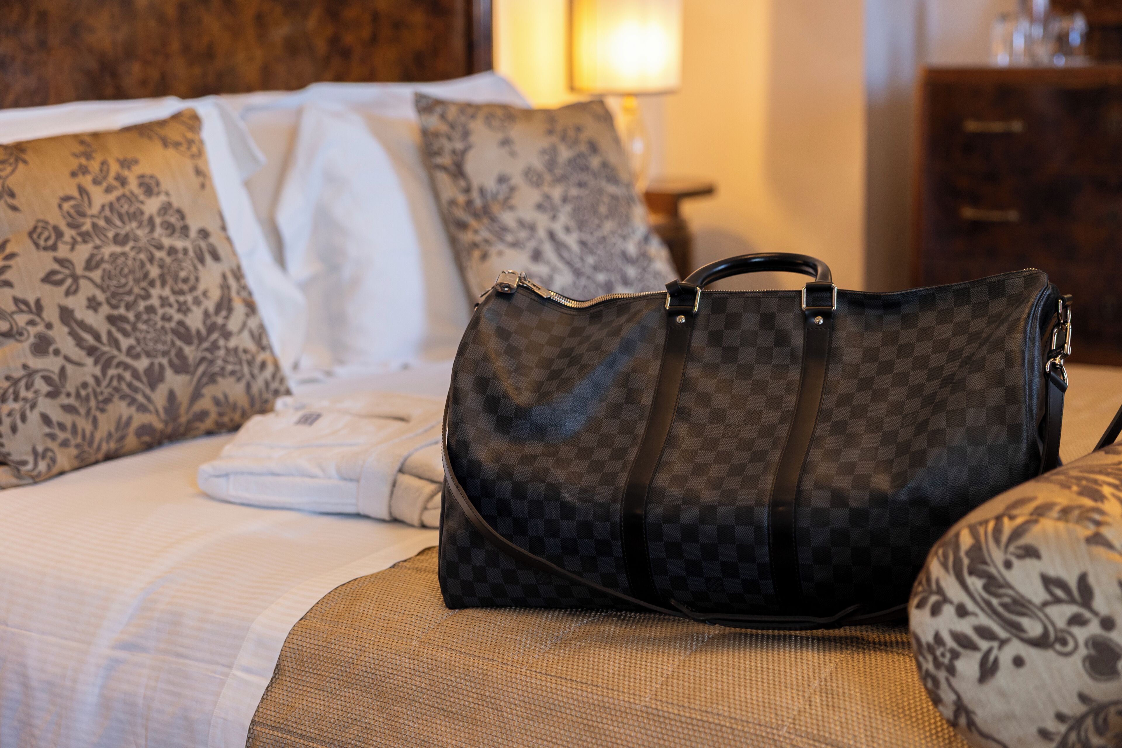 A bed with a checkered bag on it, a pillow with a floral pattern, and a lamp in the background.