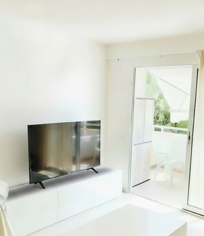 Una habitación moderna con un televisor de pantalla plana sobre un mueble blanco, y un ventanal corredero que da a un espacio exterior con sillas.