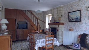 Comedor con una mesa dispuesta para cuatro, una escalera de madera, una chimenea y un televisor al fondo.