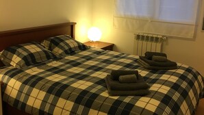 A neatly made bed with a checkered bedspread, a bedside lamp, and a radiator. - LisbonMeetU - Flat in Parque das Nações | T1 (Sacavém)
