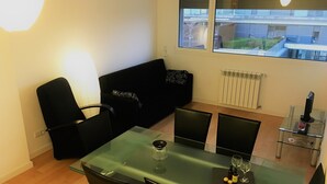 A modern living room with a glass dining table, black leather chairs, a black sofa, and a window with a view of buildings. - LisbonMeetU - Flat in Parque das Nações | T1 (Sacavém)