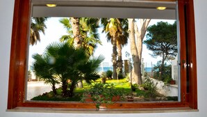 Une fenêtre donnant sur un jardin avec des palmiers et un plan d’eau.