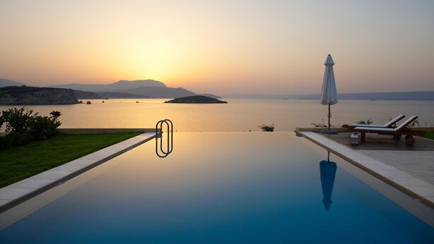 Piscina infinita com vista desimpedida do pôr do sol sobre a água, cadeiras de descanso e um guarda-sol.