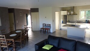 Een ruime woonkamer met een zwarte bank, een houten eettafel en stoelen. In de achtergrond is een keuken met moderne apparatuur en een groot raam zichtbaar.