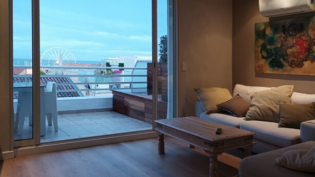 Ein modernes Wohnzimmer mit einem weißen Sofa, einem Holztisch und einem großen Fenster mit Blick auf einen Balkon. Von dort aus ist ein Riesenrad und das Meer zu sehen.