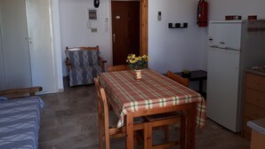 Quarto compacto com uma mesa de jantar de madeira, um sofá, um pequeno espaço com cozinha e geladeira, e um extintor de incêndio na parede.