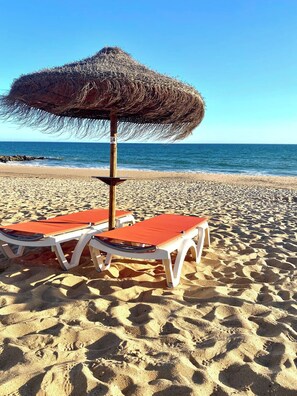 Una playa con dos sillas de playa bajo un paraguas de paja, un poste de madera y una vista despejada del océano.