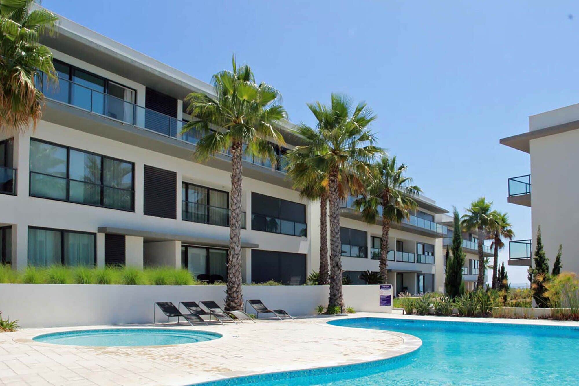 A modern hotel with a pool area, palm trees, and lounge chairs.