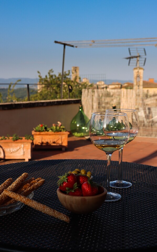 A table with two wine glasses, a bowl of strawberries, and pretzels on a rooftop terrace with a view of buildings and greenery.