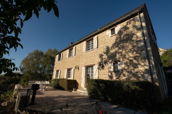 A stone building with a paved courtyard, white shutters, and a small garden area. - La Maison – Spacious Loire Cottage with Heated Pool near Chinon & Azay le Rideau (Avon-Les-Roches)