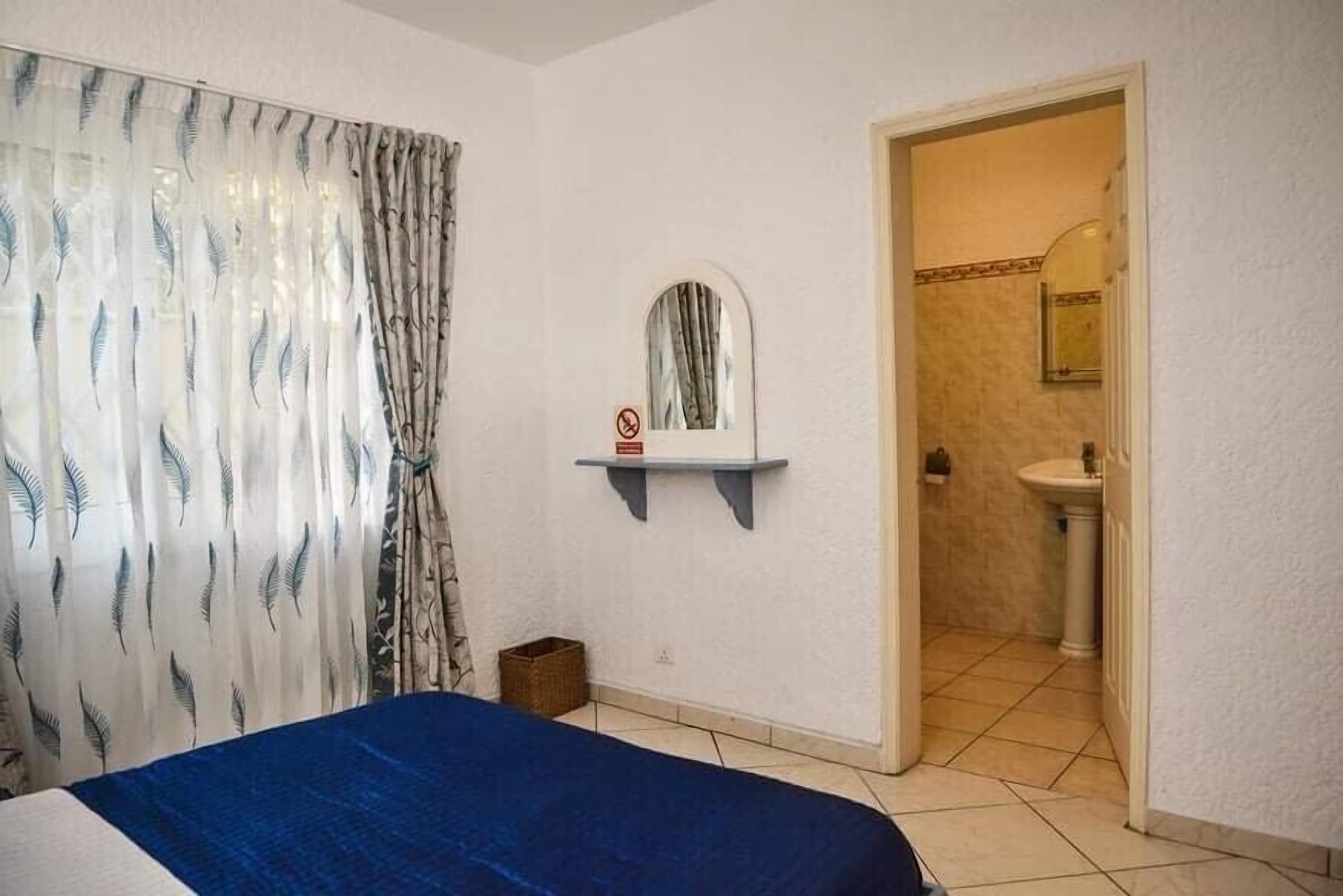 A bedroom with a bed, a window with curtains, a mirror on the wall, and a bathroom visible through an open door.