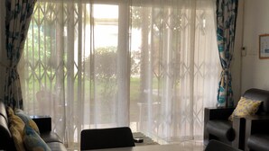 A room with a dining table, two black leather chairs, a sofa with patterned cushions, and sheer curtains with a leaf pattern. - Lovely apartment near Flic en Flac beach, the sunniest west coast of Mauritius.  (Les Orchidees)