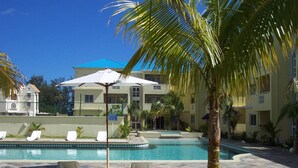 A pool area with lounge chairs and umbrellas, surrounded by palm trees and a building with a blue roof. - Lovely apartment near Flic en Flac beach, the sunniest west coast of Mauritius.  (Les Orchidees)