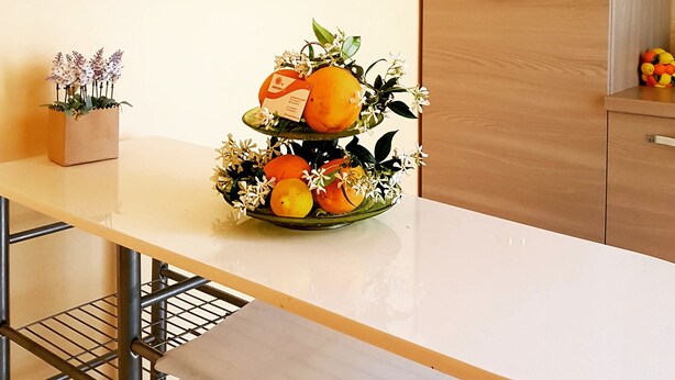 Una cocina moderna con encimera blanca, un arreglo de frutas y flores de dos niveles, y armarios de madera.