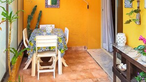 Un pequeño patio con una mesa y sillas, rodeado de plantas en macetas y una pared amarilla con azulejos decorativos y obras de arte.