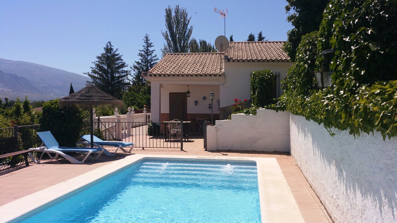 A swimming pool with lounge chairs, a small building with a tiled roof, and a mountainous backdrop.
