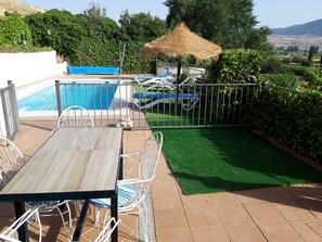 Área de piscina com mesa e cadeiras, um guarda-sol de palha e vista para a paisagem.