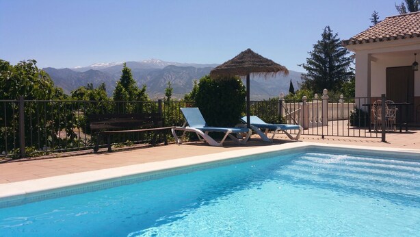 Une piscine entourée d’une clôture, avec des chaises longues et un parasol en chaume, offrant une vue sur les montagnes.