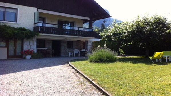 A two-story building with a balcony, a gravel driveway, and a grassy lawn with outdoor furniture. - F3 ground floor of a villa on the lake (Veyrier-Du-Lac)