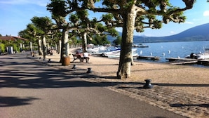 Une promenade au bord du lac, bordée d’arbres, avec des bancs et des bateaux amarrés sur la rive.