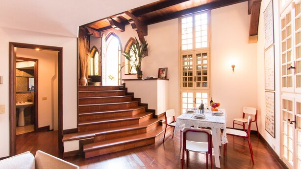 Un comedor con una escalera de madera, una mesa dispuesta para dos personas y vista a una cocina al fondo.