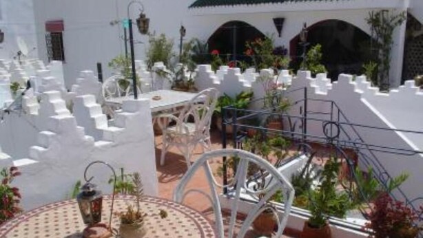 Een terras op het dak met witte muren, een ronde tafel met een tafelkleed met patroon, en plantenpotten.