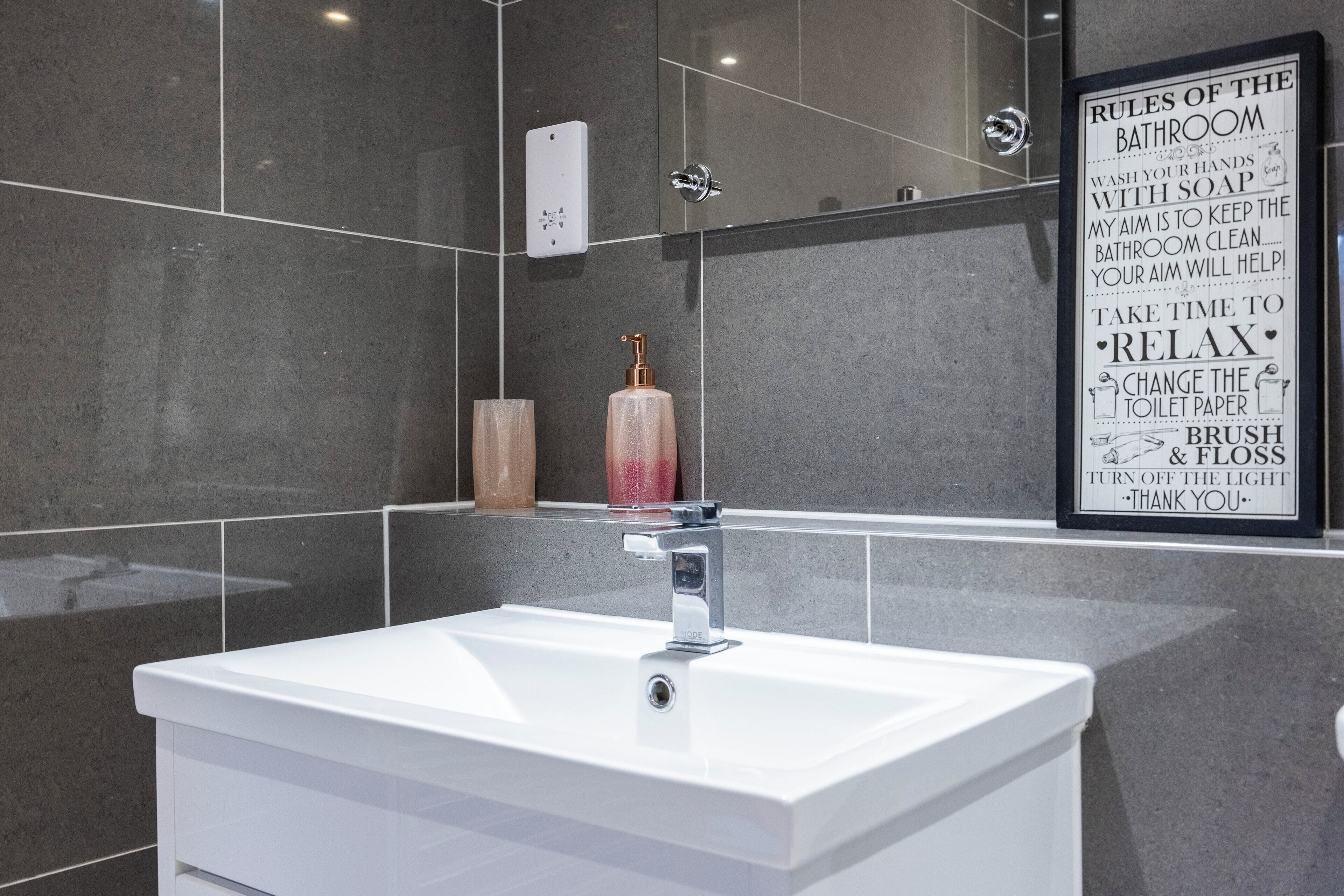 Un baño moderno con un lavabo blanco, un dispensador de jabón para manos y un cartel con normas del baño.